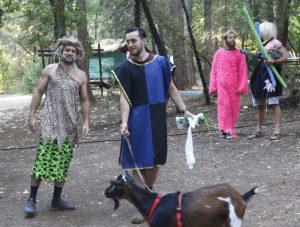 Four adults in playful, colorful costumes stand in a wooded outdoor area. One man holds a goat on a leash. Two people are dressed in caveman outfits, while two others wear whimsical pink and blue costumes and carry props.