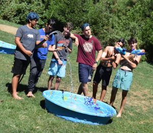 Six teenage boys stand around a small kiddie pool on grass. Some are holding water guns, and one is spraying water into the pool. They appear to be having fun outdoors on a sunny day.