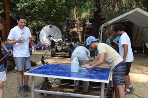 Several young people are gathered outdoors around a blue ping pong table, with one person cleaning it using a sponge and jug of soapy water. Trees and tents are visible in the background.