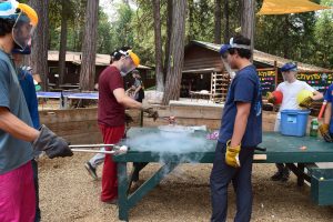 Several people wearing gloves and face shields conduct a science experiment with dry ice at a green table outdoors, surrounded by trees and wooden buildings, with smoke rising from a bowl.