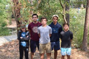 Five young people stand outdoors in a wooded area, facing the camera. Four boys and one girl, wearing casual summer clothes, stand among trees and dry leaves, with neutral expressions.