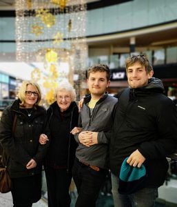 Four people stand close together, smiling, in a shopping mall decorated with hanging golden and white ornaments. Two women and two men are dressed warmly, and one man holds a blue hat.