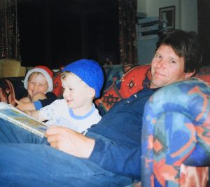A man sits on a colorful couch with two smiling young boys. One boy wears a Santa hat, the other a blue knit hat. The man holds a book, and they appear to be reading together in a cozy living room.
