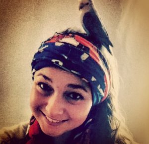 A smiling person wearing a colorful scarf on their head has a small bird perched on top of the scarf. The image has a warm, vintage filter.
