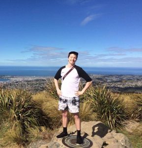 A person stands smiling on a rocky hilltop, hands on hips, with a city, coastline, and ocean visible in the background under a clear blue sky. Grassy plants surround the spot.