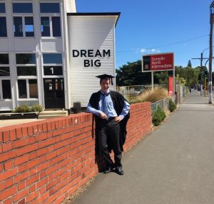 A person wearing graduation attire stands by a red brick wall in front of a building with large letters that read DREAM BIG. A school sign for Dunedin North Intermediate is visible in the background.