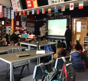 A classroom with students sitting on the floor facing a whiteboard, where a teacher is pointing at the screen and another adult stands nearby. Desks, chairs, and international flags hang from the ceiling.
