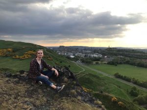 A person sits on a rocky hilltop with green hills and a cityscape in the background under a cloudy sky, possibly near sunset. Yellow flowers dot the hills and a road winds through the landscape below.