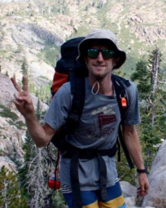 A person wearing sunglasses, a gray hat, and a backpack flashes a peace sign while hiking in a mountainous, forested area. The sun is shining and trees and rocky hills are visible in the background.