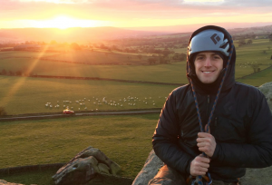 A smiling person in climbing gear sits on a rocky ledge at sunset, with rolling green fields, grazing sheep, and a golden sun on the horizon in the background.
