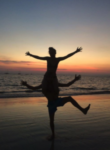 Two people are silhouetted against a colorful sunset on the beach; one is sitting on the others shoulders with arms outstretched, while the person below balances on one leg with arms raised.