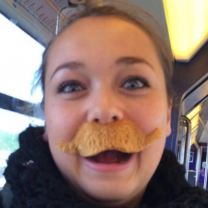 A young woman with blue eyes and light brown hair is smiling widely on public transportation, wearing a large, fake, bushy, brown mustache. The background shows bus or train seats and windows.