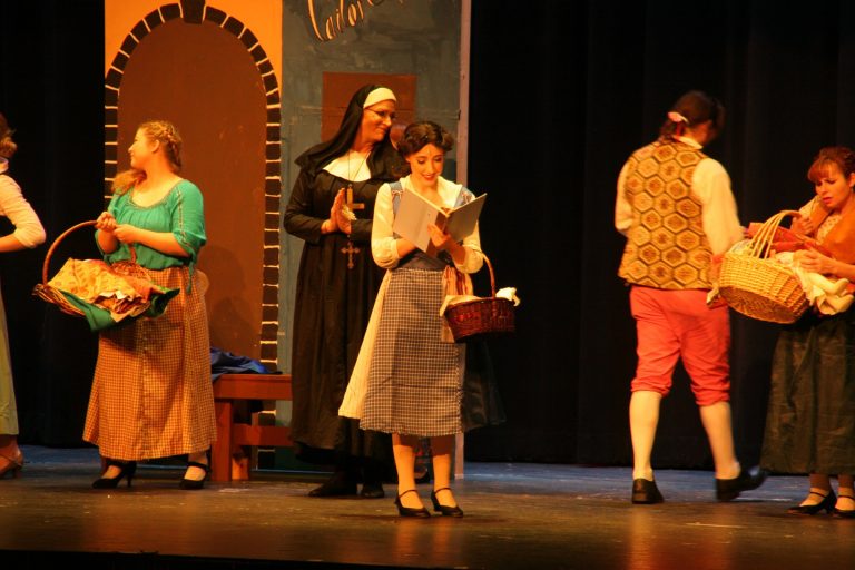 Bailey standing mid stage reading book as Belle while townspeople move around her
