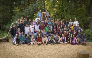 A large, diverse group of people pose together outdoors in front of green trees, smiling and laughing for a photo. Some are sitting while others stand behind, with a few stacked in playful poses on a small wooden ladder—perfect for summer camp staff bios.