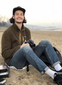 A young man with long hair wearing a cap, brown fleece jacket, jeans, and white socks sits on a sandy beach, smiling and holding a camera. A backpack and some belongings are beside him. The background is misty with distant trees.