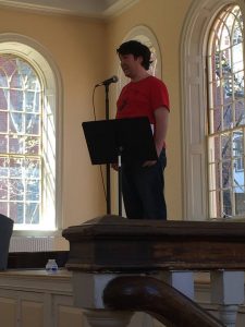 A man in a red shirt stands at a microphone on a stage, reading from a music stand in a sunlit room with large arched windows. A water bottle sits on the stage beside him.