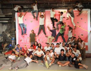 A large group of smiling people poses together indoors, some sitting on the floor and others climbing on a wall with colorful holds. A pink banner with AIRPLAY is in the background. The mood is joyful and energetic.