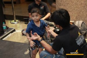 A young boy wearing a blue shirt and climbing harness listens to an instructor, who gestures with his hands. Both are indoors, with climbing gear and a Natura Adventure Services logo visible.
