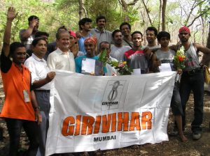 A group of smiling people outdoors hold a large white banner that says GIRIVIHAR MUMBAI. Some hold certificates and flowers, and trees are visible in the background. It appears to be a celebratory event.
