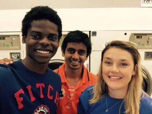 Three young adults smile for a selfie in a laundromat, standing in front of washing machines. One wears a blue FITCH shirt, another an orange shirt, and the third a blue shirt with blonde hair.