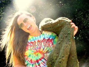A smiling person in sunglasses and a colorful tie-dye shirt holds a large alligator sculpture outdoors, with sunlight streaming in from behind.