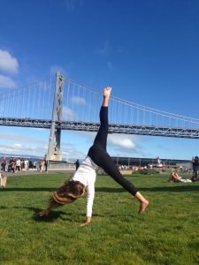 A woman performs a cartwheel on grass near a large suspension bridge, with people relaxing and walking in the background under a bright blue sky.