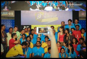 A large group of people in colorful shirts pose and smile for a group photo at the IFSC Climbing World Cup event, while someone in the foreground takes a photo with a smartphone.