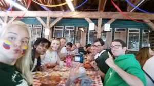 A group of young adults sit at a long table with checkered tablecloth, smiling and posing for a selfie. Some have their faces painted with colorful flags. The setting is a rustic hall decorated with streamers.