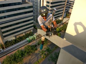 A worker wearing a helmet, gloves, and safety gear is out on a high beam of a tall building, using tools to secure a cable, with city buildings and a street far below.