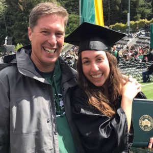 A young woman in a graduation cap and gown smiles while holding her diploma next to a man, both standing outdoors at a graduation ceremony. Rows of chairs and people are visible in the background.