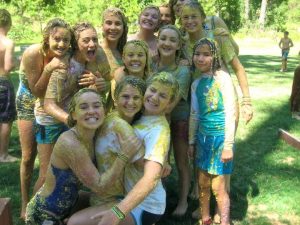 A group of smiling kids and teens, covered in yellow paint, huddle together outdoors on green grass on a sunny day, enjoying what seems to be a fun, messy activity. Trees and more people are visible in the background.