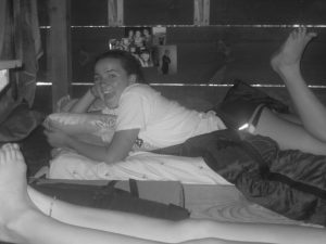 A girl lies on a bed in a wooden room, smiling with her mouth open and holding a pillow. Her legs are bent, and another person’s legs are visible in the foreground. Photos are taped to the wall behind her.