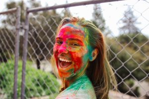 A person with long hair smiles joyfully, their face covered in bright, multicolored powder. They stand outdoors in front of a chain-link fence, with trees visible in the background.