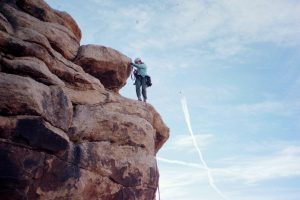 A person in climbing gear scales a large rocky formation, reaching toward the top against a blue sky with thin clouds and jet trails.