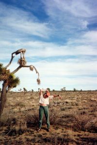 A person stands in a dry desert landscape near a Joshua tree, mimicking the trees bent arm-like branches with their own arms raised and bent, under a mostly cloudy blue sky.