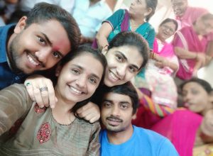 Four people are smiling and posing closely for a selfie, while several others in colorful traditional clothing sit and chat in the blurred background, creating a cheerful, lively atmosphere.