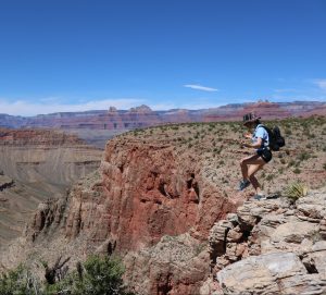 A person wearing a hat, blue shirt, and backpack stands near the edge of a rocky cliff overlooking the vast, layered red rock formations of the Grand Canyon under a clear blue sky.