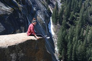 A person in a red plaid shirt and striped pants sits on the edge of a large cliff, legs dangling, with a steep drop and forested valley in the background.
