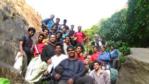 A group of around 25 people pose together outdoors on a rocky trail surrounded by greenery, some holding climbing gear and ropes, all smiling at the camera.