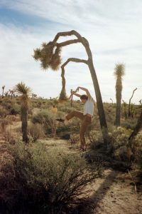 A person in casual clothes poses playfully, mimicking the shape of a bent Joshua tree behind them in a desert landscape under a partly cloudy sky.