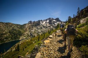 Two hikers with backpacks walk along a rocky mountain trail with a scenic view of jagged peaks, a lake, and lush green forests under a clear blue sky.