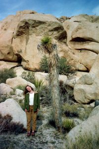 A woman in a green jacket and tan pants stands tilting her head beside a drooping, shaggy-looking plant in a rocky desert landscape with large boulders and sparse vegetation.