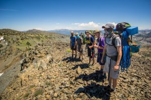 A group of six hikers wearing backpacks and hats stand on a rocky mountain ridge under a clear blue sky, with distant mountains visible in the background.