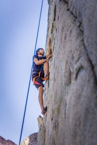 A person wearing climbing gear and a helmet is scaling a steep rock face, secured with a blue rope, against a clear blue sky.
