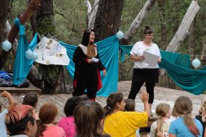 Two women perform on an outdoor stage in front of children; one wears a bat costume, the other holds a gift bag. Blue fabric and drawings decorate the stage, with trees and forest in the background.