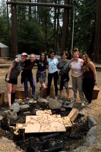 Seven smiling teens stand arm-in-arm around a campfire ring in a wooded outdoor camp. Behind them are tall trees, tents, and a wooden sign with a painted tree and symbols resting on the firepit.