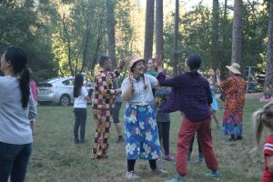 A group of people dance outdoors in a grassy, wooded area. Some wear colorful, patterned outfits, and two women in the center face each other, clapping hands and smiling. Cars are parked in the background.