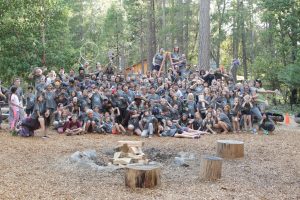 A large group of children and adults pose together outdoors in a wooded area, many smiling and making playful gestures around a campfire ring with logs and rocks. Trees and a cabin are visible in the background.