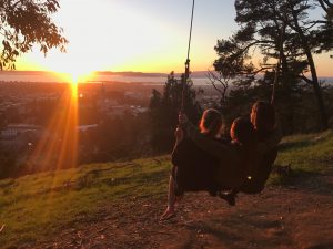 Three people sit together on a swing, facing a vibrant sunset over a city and body of water, with trees and grass around them, creating a peaceful and scenic view.