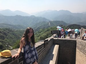 A woman in a blue dress smiles while standing on the Great Wall of China, with green mountains and a group of tourists under umbrellas in the background on a sunny day.